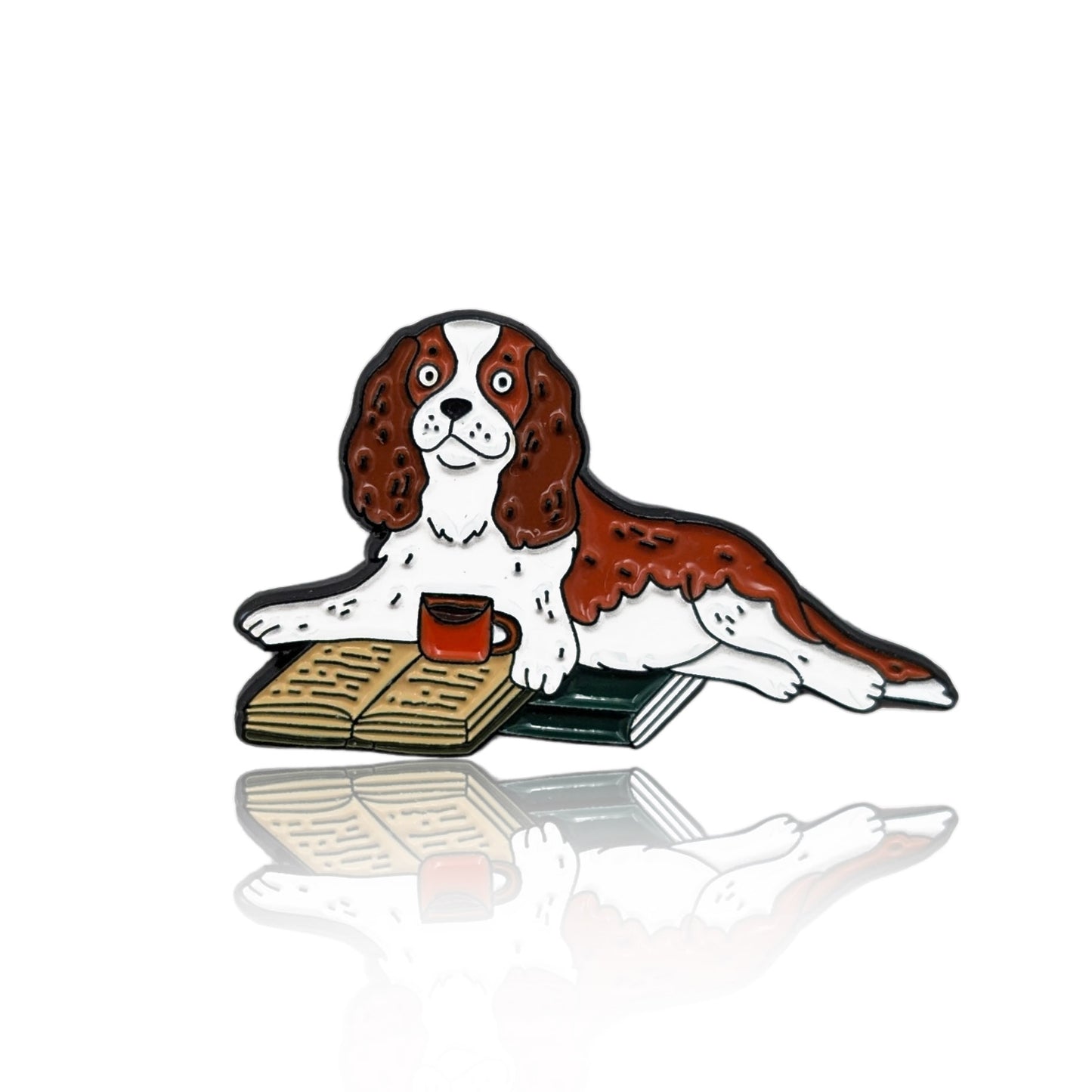 Enamel pin of a cartoon Cavalier dog lying on books with a red mug and an open book