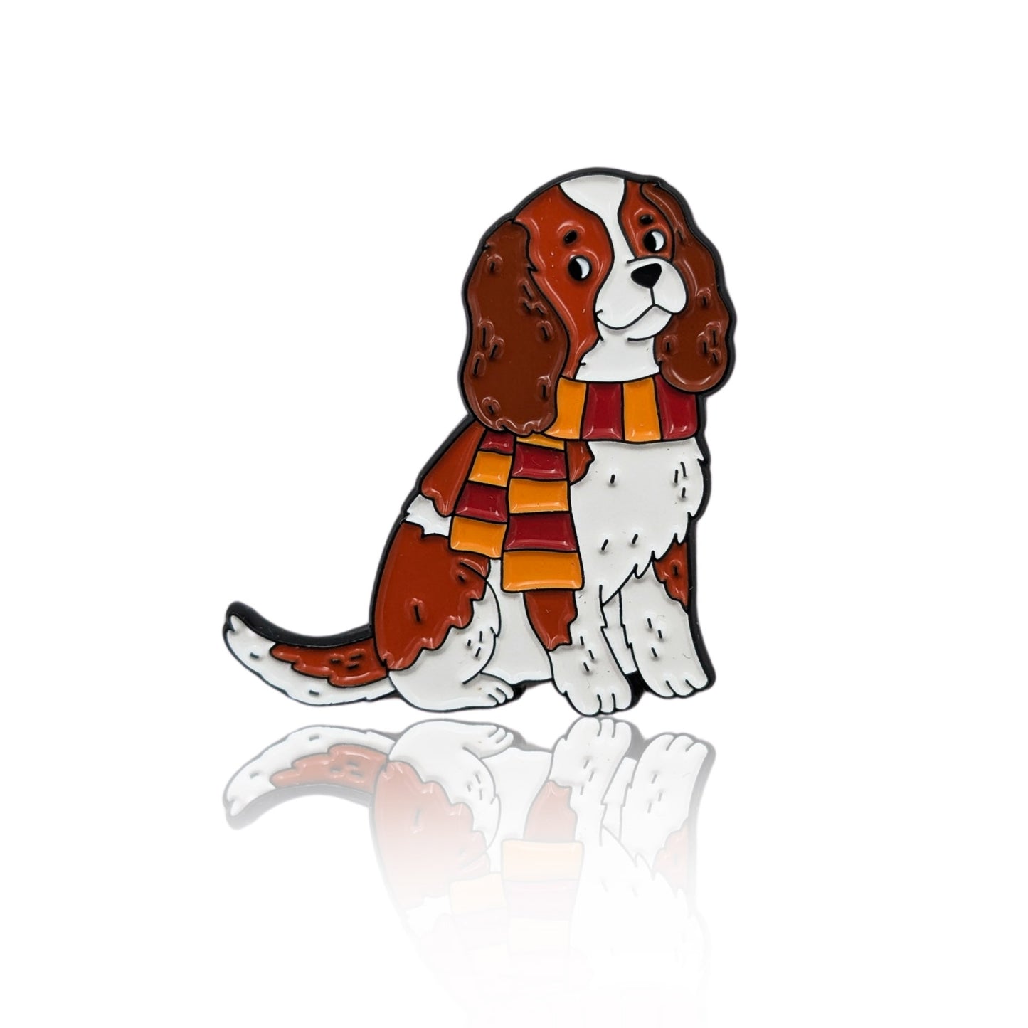 This 32 mm wide enamel pin features a cartoon-style Cavalier King Charles Spaniel sitting down and wearing a red and gold striped scarf. Designed by PINBOX artists in 2025. A cozy and stylish pin for backpacks, jackets, or autumn outfits.