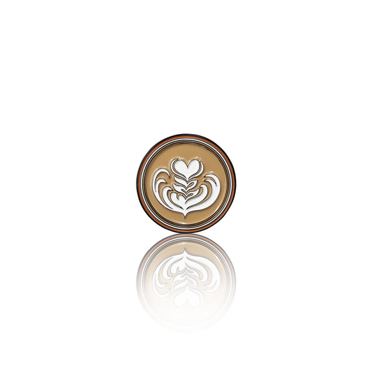 Round enamel pin on a white background with a creamy coffee base and a white latte art heart design, shown with its reflection.