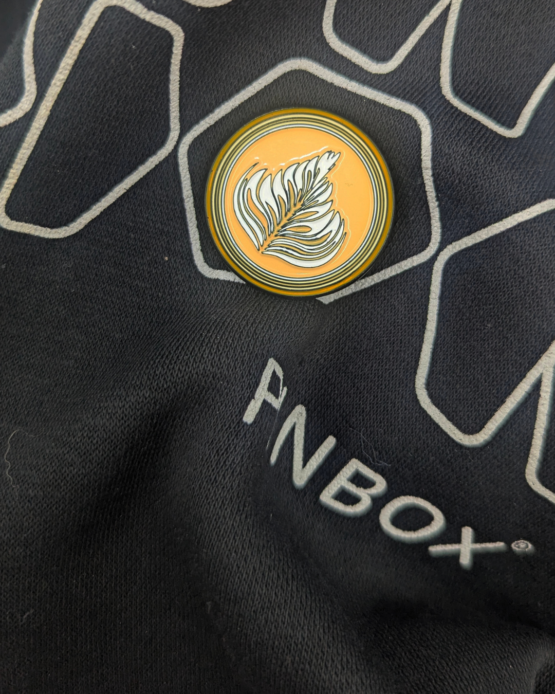 Round enamel pin with a golden latte background and white leaf art, fastened to black clothing with PINBOX branding.