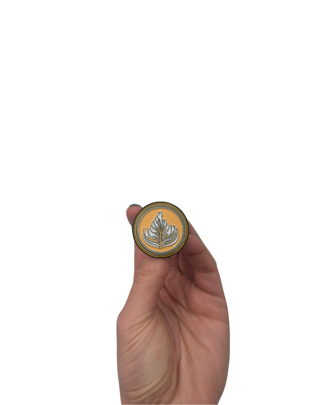 Hand holding a round enamel pin designed like a coffee latte with a white leaf art pattern on a golden base.