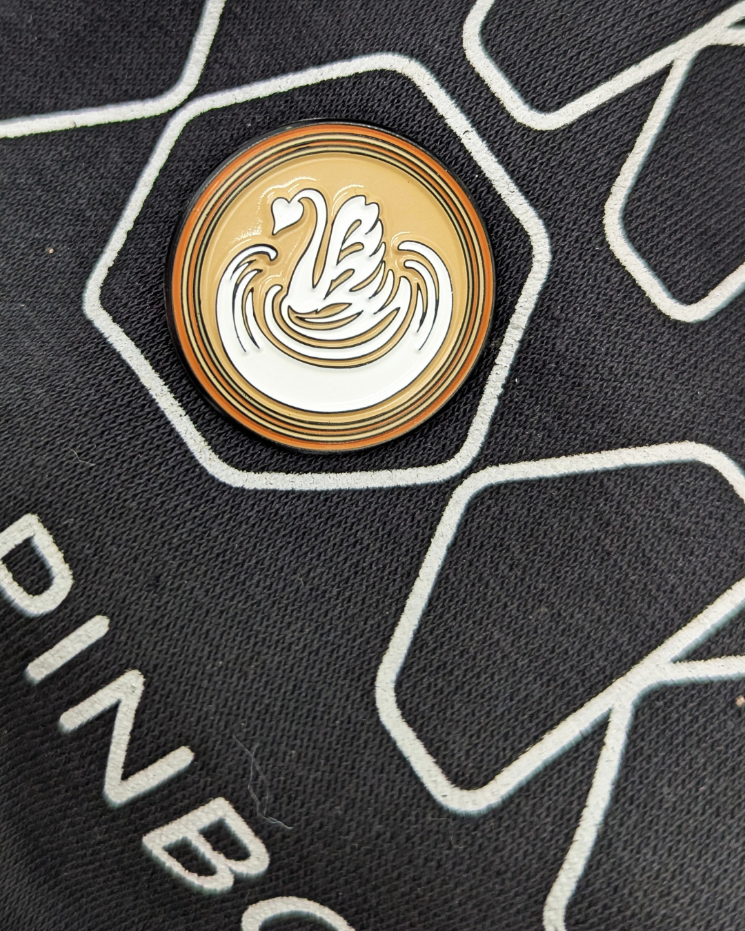 Enamel pin with a coffee base and white swan latte art design, fastened onto black clothing with PINBOX branding visible.