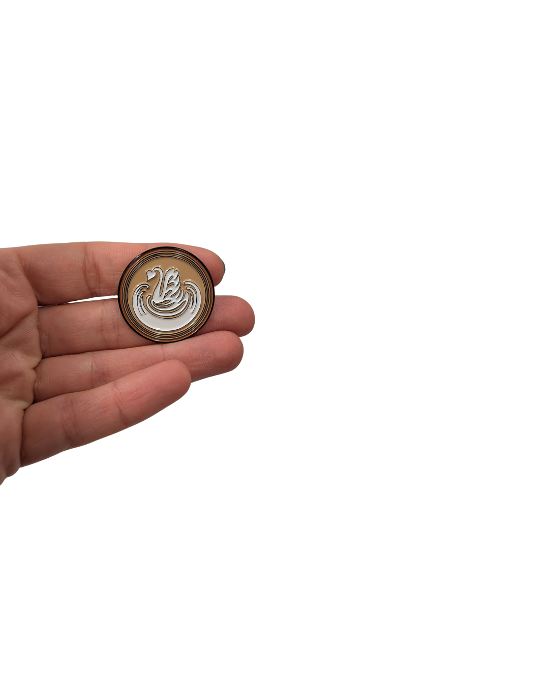 Hand holding a round enamel pin featuring a golden coffee-colored base with a white swan-shaped latte art design.
