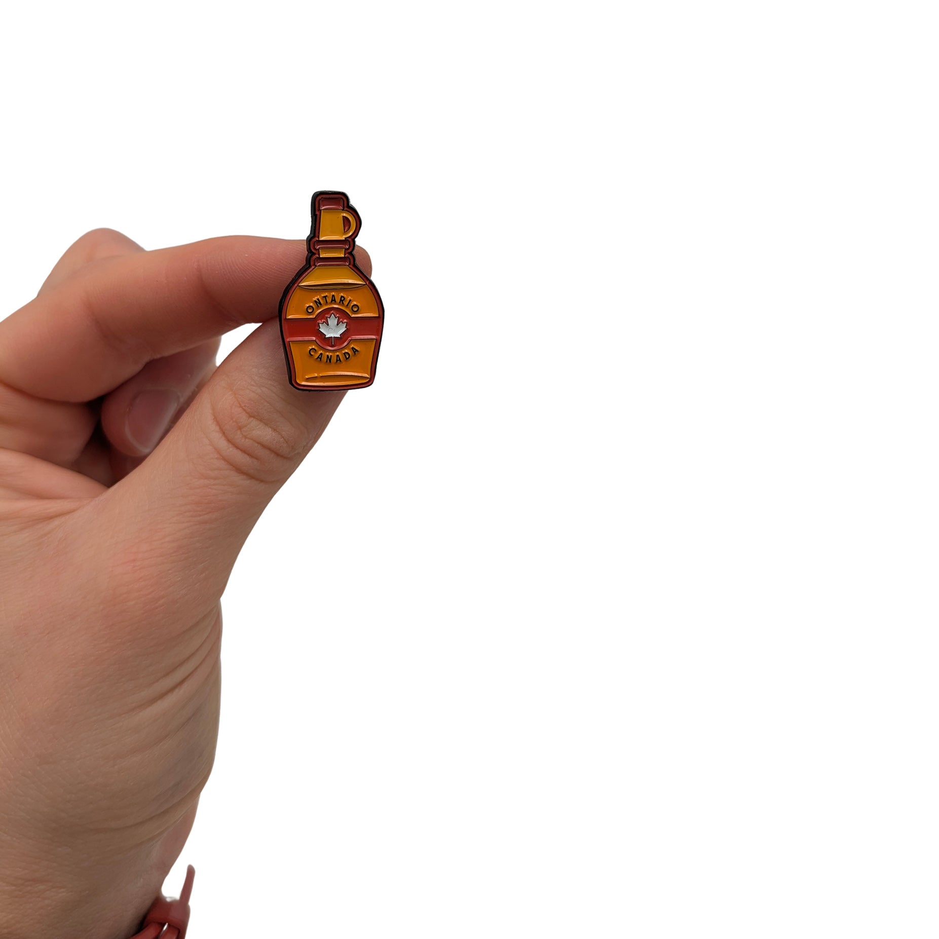 Maple syrup bottle enamel pin held in hand, featuring amber and red colors with a maple leaf detail