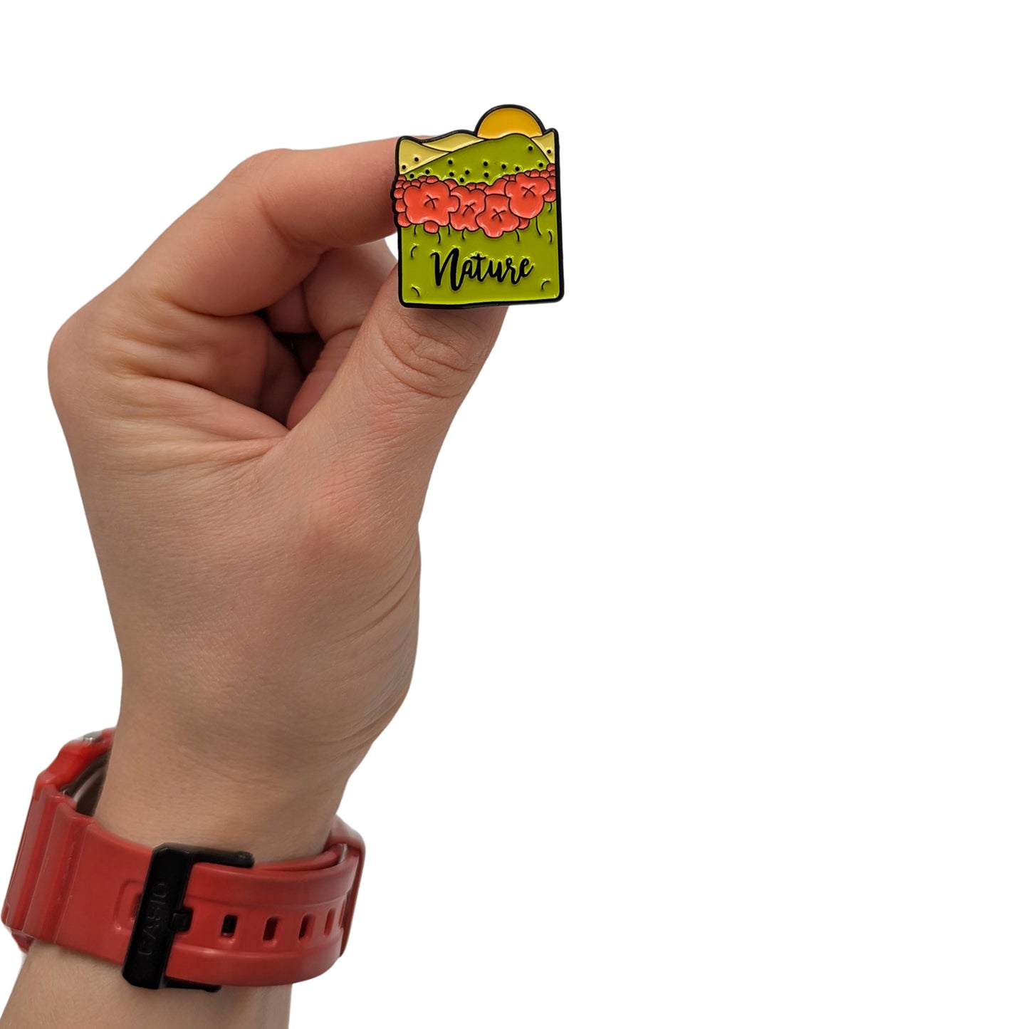 Nature landscape enamel pin held in hand, featuring flowers, green hills, and a sun with the word “Nature”