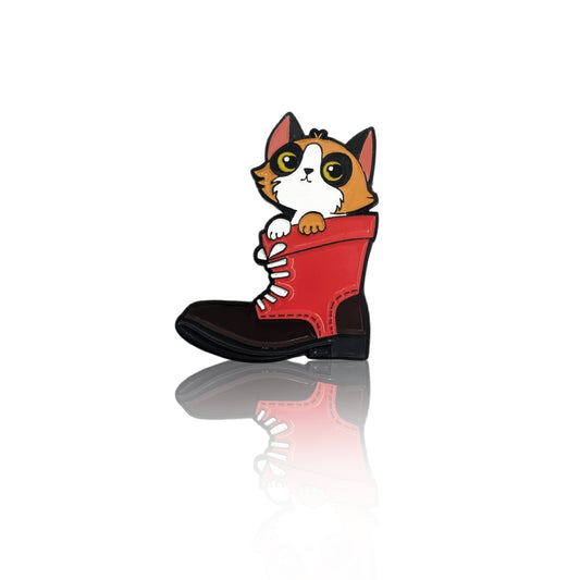 enamel pin showing an orange and white cat sitting inside a red lace-up boot on white background